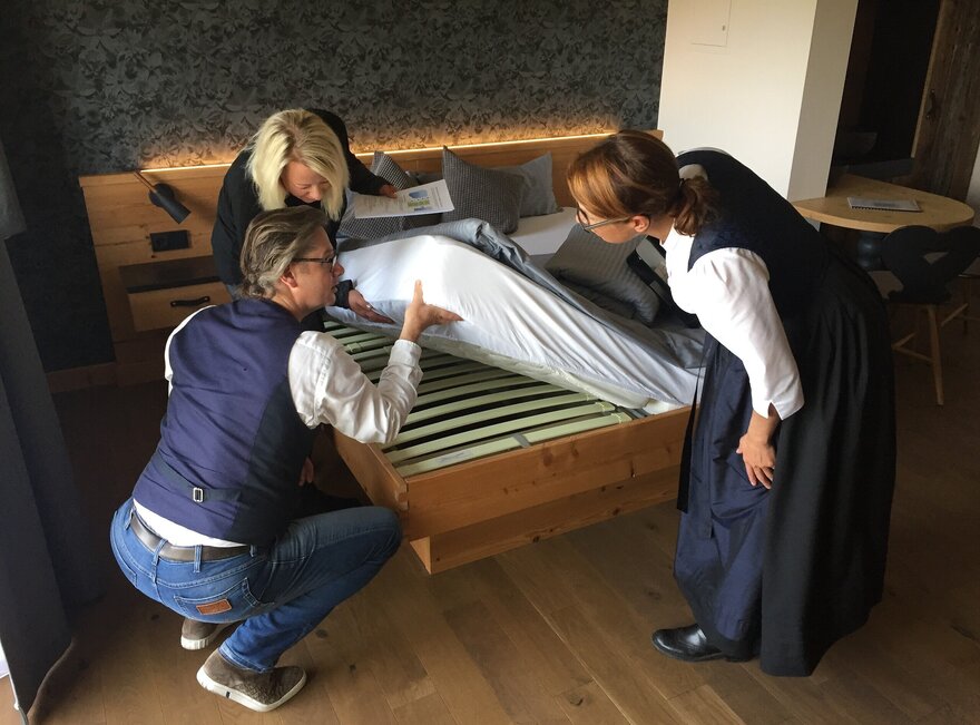 Das Bild zeigt drei Personen, die die Matratze eine Bettes hochheben und den darunterliegenden Lattenrost betrachten. Eine Frau trägt ein dunkles Dirndl. Eine Frau hat Papiere in der Hand. Ein Mann in Jeans, weißem Hemd und Weste ist in die Hocke gegangen.