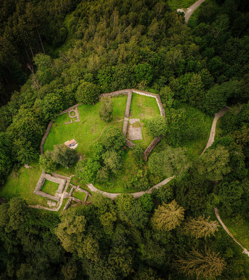 Das Bild zeigt die Mauerreste einer Burg. Das Foto ist aus der Vogelperspektive aufgenommen. Aus diesem Blickwinkel wirken die Mauerreste wie ein Grundriss, des ehemaligen Burggebäudes. Die Ruine steht auf einer Anhöhe und ist umgeben von dichtem Nadelwald. Es handelt sich um die Ruine der einstigen Burg Hopfen.