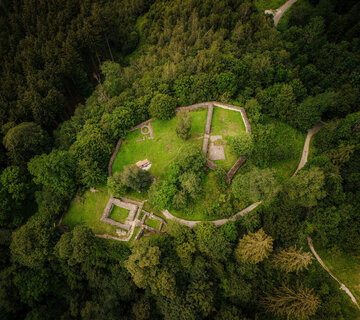 Das Bild zeigt die Mauerreste einer Burg. Das Foto ist aus der Vogelperspektive aufgenommen. Aus diesem Blickwinkel wirken die Mauerreste wie ein Grundriss, des ehemaligen Burggebäudes. Die Ruine steht auf einer Anhöhe und ist umgeben von dichtem Nadelwald. Es handelt sich um die Ruine der einstigen Burg Hopfen.