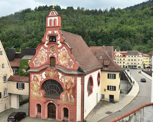Das Foto zeigt die reich verzierte und bemalte Fassade der Spitalkirche in Füssen. Unterschiedliche Heilige sind dort abgebildet.  Im Hintergrund sieht man den beginnenden Bergwald. Die Kirche ist umrahmt von weiteren eher schlichten Gebäuden. Im Vordergrund verläuft eine Straße. 