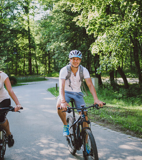 Das Bild zeigt einen Mann und eine Frau beim Fahrradfahren. Sie fahren auf E-Bikes, tragen Freizeitkleidung und Helme. Sie fahren auf den Betrachter zu auf einer asphaltierten Straße, die von Wäldern umgeben ist. Beide lächeln. Die Sonne scheint.