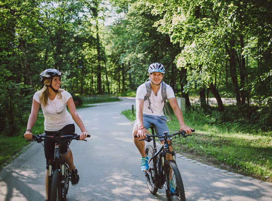Das Bild zeigt einen Mann und eine Frau beim Fahrradfahren. Sie fahren auf E-Bikes, tragen Freizeitkleidung und Helme. Sie fahren auf den Betrachter zu auf einer asphaltierten Straße, die von Wäldern umgeben ist. Beide lächeln. Die Sonne scheint.
