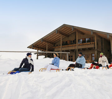 Das Bild zeigt fünf Personen, die je auf einem Holzschlitten sitzen. Die Schlitten stehen hintereinander vor einem Holzhaus. Alle tragen Winterkleidung und Mützen. Alle lächeln. Die Landschaft ringsum ist tief verschneit. Die Sonne scheint.