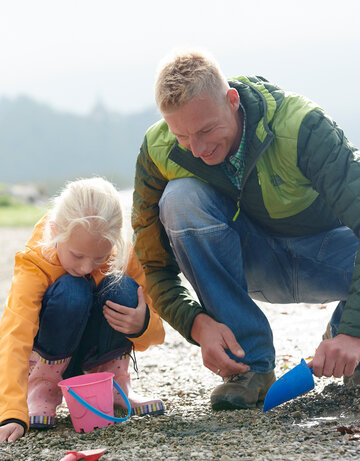 Das Bild zeigt einen Mann und ein Mädchen. Sie knien beide an einem steinigen Seeufer. Das Wetter ist sehr nebelig und sie tragen Regenkleidung. Sie spielen mit einer kleinen Plastikschaufel und einem Eimer in den Steinen. 