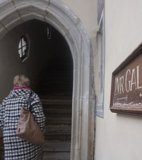 Das Bild zeigt eine Frau in einem karierten Mantel. Sie hat eine große braune Handtasche über der Schulter und dreht dem Betrachter den Rücken zu. Sie schreitet durch ein spitz zulaufendes Bogenportal. An das ein steinernes Treppenhaus anschließt. 