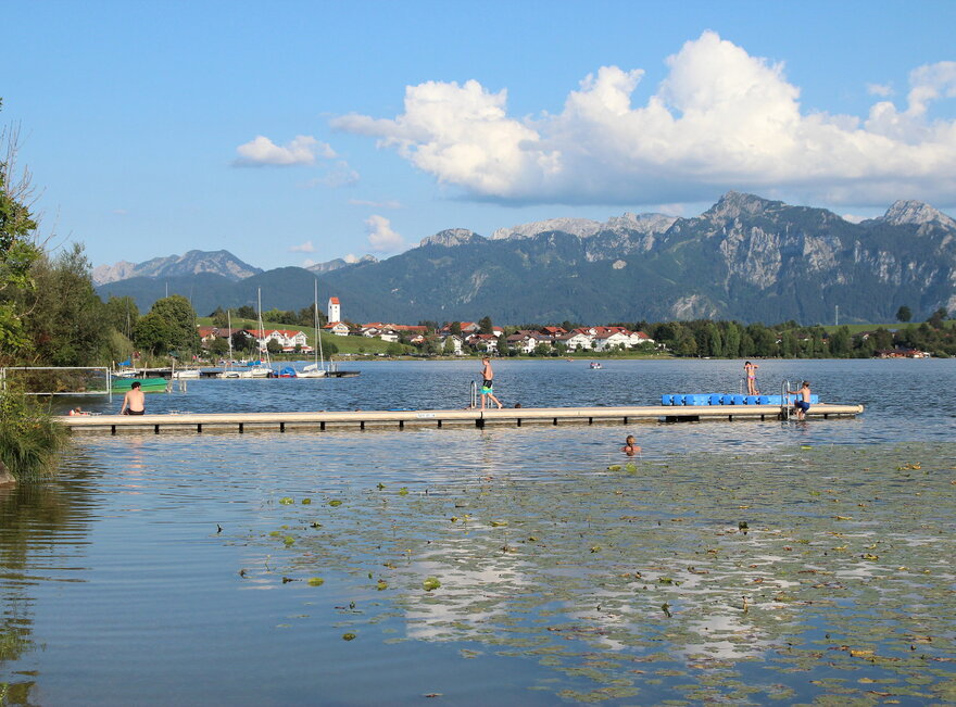 Das Bild zeigt einen See, auf den vom linken Bildrand aus ein hölzerner Steg ins Wasser führt. Auf dem Steg sitzen und laufen Menschen. Im Wasser schwimmt eine langhaarige Person. Im Vordergrund sind unzählige Blätter uns Knospen einer gelben Seerose zu sehen. Die Knospen sind noch nicht geöffnet. Im Hintergrund erheben sich die bewaldeten Berge. Davor liegt, am Ufer des Sees, eine kleine Ortschaft mit Kirchturm und Häusern mit roten Dächern. Der Himmel ist blau. Die Sonne scheint.