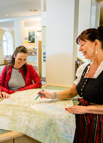 Das Bild zeigt einen Mann und zwei Frauen, die an einem Informationstresen stehen. Der Mann und die Frau, die hinter dem Tresen stehen, tragen Freizeitkleidung. Die Dame auf der anderen Seite trägt ein Dirndl mit einer grün-roten Schürze. Alle lächeln. Die Frau im Dirndl deutet mit einem Stift auf eine Karte, die vor den Personen auf dem Tresen liegt. Alle lächeln.