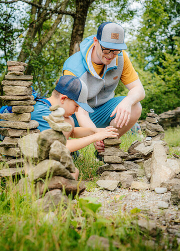  Das Bild zeigt einen Moment der Verbindung zwischen einem Mann und einem jungen Jungen in einer ruhigen Landschaft. Der Mann, der ein blaues Baseball-Cap und eine gelbe Jacke trägt, ist neben dem Jungen auf den Knien, der ein blaues Baseball-Cap und ein blaues Hemd trägt. Sie sind beide in die spielerische Aktivität des Steine stapeln vertieft. Die Steine, die in Größe und Form variieren, liegen um sie verteilt und bilden einen improvisierten Stein-Turm. Der Hintergrund ist mit üppiger Vegetation gefüllt, was eine natürliche und friedliche Kulisse für ihre Aktivität bildet. Das Bild strahlt eine Atmosphäre von Spaß, Lernen und Verbindung zwischen den beiden Individuen aus.