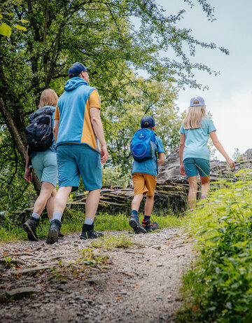 Das Bild zeigt eine Familie, bestehend aus einer Frau, einem Mann, einem Jungen und einem Mädchen, die einen Wanderweg nach oben gehen. Sie tragen sportliche Freizeitkleidung. Im Hintergrund ist die Mauer einer Ruine zu sehen. Ringsum wachsen Bäume, Büsche und Gräser. Der Himmel ist blau. Die Sonne scheint.