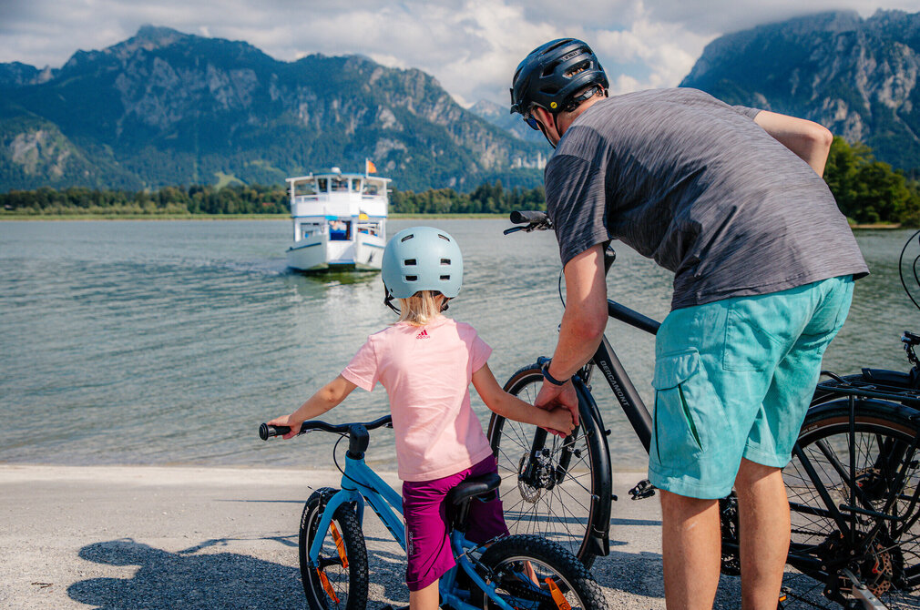 Das Bild zeigt einen friedlichen Moment auf einem See, wo zwei Frauen eine Stand-Up-Paddling-Session genießen. Die Frau in der Vordergrund steht auf dem SUP-Board, hält einen Paddel und scheint in Bewegung zu sein. Sie trägt eine pinkfarbene Schwimmweste, die gegen das ruhige Wasser kontrastiert. Die zweite Frau sitzt auf dem SUP-Board, ebenfalls mit einer Schwimmweste ausgestattet, und blickt in Richtung Kamera. Beide Frauen tragen Badeanzüge, die für die Wassersportaktivität geeignet sind. Der See wird von Bergen umgeben, was die Kulisse malerisch macht. Der Himmel über der Szene ist von Wolken erfüllt, was auf einen bedeckten Tag hindeutet. Die Gesamtatmosphäre des Bildes ist ausgestattet mit Entspannung und Genuss der Natur.