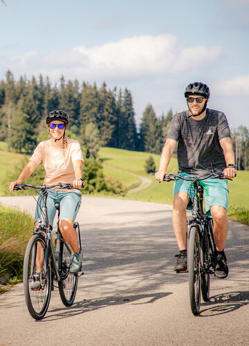 Das Bild zeigt einen Mann und eine Frau beim Fahrradfahren. Beide tragen Freizeitkleidung und einen Helm. Sie fahren auf den Betrachter zu. Beide lächeln und tragen Sonnenbrillen. Die Straße, auf der sie fahren ist asphaltiert. Wiesen und Wälder säumen die Straße. Am Straßenrand ist eine Wegkreuz aus Metall und eine Holzbank zu sehen.  Im Hintergrund erheben bewaldete Hügel. Der Himmel ist blau. Die Sonne scheint. 