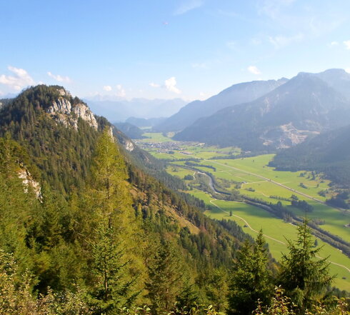 Man blickt von einer Anhöhe in ein grünes Tal, durch das ein Fluss fliest. Das Tal ist flankiert von hohen Bergen und bewaldeten Anhöhen. Der Himmel ist blau. Die Sonne scheint. 