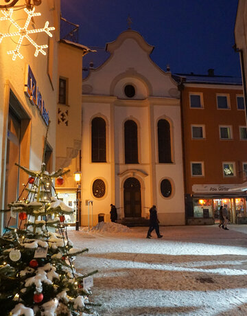 Das Bild fängt eine friedliche Nachtszene in einer europäischen Stadt ein. Der Fokus liegt auf einem wunderschön dekorierten Weihnachtsbaum, der in der Vordergrund steht. Der Baum ist mit funkelndem Lichtern und Ornamenten geschmückt, die eine warme Glut auf die umgebende Fläche werfen.  Im Hintergrund steht eine große Kirche mit einer weißen Fassade und einem roten Dach majestätisch. Die architektonischen Details der Kirche sind deutlich sichtbar und fügen dem Charme der Szene hinzu.  Die Straße, wo der Baum steht, ist von Gebäuden gesäumt, deren Fenster mit dem weichen Licht der Kirche leuchten. Einige Menschen können gesehen werden, die auf der Straße gehen, fügen einen Hauch von Leben zu dem sonst stillen Bild hinzu.  Der Himmel über ist ein tiefes Blau, das eine eindrucksvolle Kulisse für die festlichen Dekorationen bietet. Die allgemeine Atmosphäre des Bildes ist eine der Ruhe und Gelassenheit, die die Magie der Weihnachtszeit einfängt.