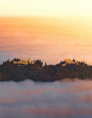 Das Bild fängt eine atemberaubende Aussicht einer Burg ein, die auf einem Hügel thront und von einem üppigen Wald umgeben ist. Die Burg, mit ihrer majestätischen Architektur, hebt sich vor dem Hintergrund des Himmels ab. Der Himmel ist in Tönen von Orange und Rosa gemalt, was darauf hindeutet, dass die Sonne gerade untergeht und die ganze Szene in ein warmes Licht taucht. Der auf dem Hügel liegende Burgberg ist mit Nebel bedeckt, was der Szene etwas Mystisches verleiht.