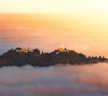 Das Bild fängt eine atemberaubende Aussicht einer Burg ein, die auf einem Hügel thront und von einem üppigen Wald umgeben ist. Die Burg, mit ihrer majestätischen Architektur, hebt sich vor dem Hintergrund des Himmels ab. Der Himmel ist in Tönen von Orange und Rosa gemalt, was darauf hindeutet, dass die Sonne gerade untergeht und die ganze Szene in ein warmes Licht taucht. Der auf dem Hügel liegende Burgberg ist mit Nebel bedeckt, was der Szene etwas Mystisches verleiht.