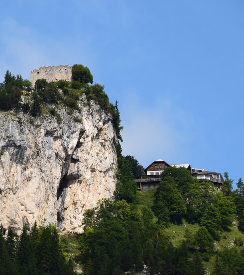 Das Bild zeigt einen atemberaubenden Ausblick auf eine Burg-ähnliche Struktur, die sich auf einem steilen Felsvorsprung befindet. Die Burg in Stein erbaut, steht majestätisch vor einem klaren blauen Himmel. Der Felsvorsprung selbst ist ein Spektakel, mit üppigem grünen Laubwerk und Büschen, die eine Naturpracht hinzufügen. Die Lage der Burg auf dem Felsvorsprung bietet einen Vorteilspunkt mit einer Panoramasicht auf das umliegende Landesinnere. Das Bild ist ein Zeugnis der architektonischen Geschicklichkeit der Vergangenheit und steht kraftvoll unter den natürlichen Elementen.