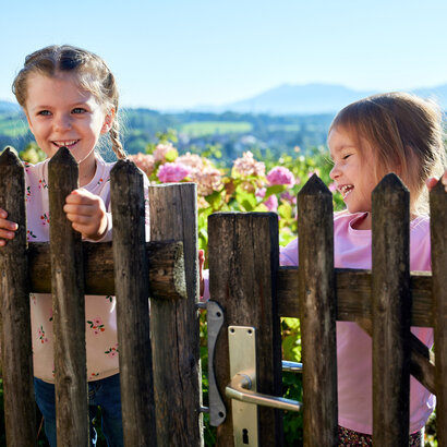 Das Bild erfasst einen herzerfrischenden Moment von zwei jungen Mädchen, beide gekleidet in Pink, die vor einem hölzernen Zaun stehen. Das Mädchen auf der linken Seite hält sich am Zaun fest, ihr Gesicht erstrahlt in einem fröhlichen Lächeln. Ihre Begleiterin auf der rechten Seite lehnt sich an den Zaun, auch sie lächelt. Sie sind von einer üppigen Gartenlandschaft umgeben, gefüllt mit lebhaften Blumen und üppigem Grün. In der Ferne sind imposante Berge zu sehen, die der Szene ein Gefühl von Wunder und Abenteuer vermitteln. Die pinkfarbenen Outfits der Mädchen kontrastieren wunderschön mit den natürlichen Farben des Gartens und der Berge, was das Bild noch faszinierender macht.