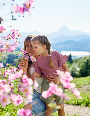Die Frau und das Kind stehen in einem Garten, der voll von blühenden Pflanzen ist. Die Frau hält einen Strauß Blumen in der Hand und schneidet eine Blume. Das Kind, das auf dem Rücken der Frau sitzt, schaut in die Blumen. Beide sind in einem Moment der Natur zugewandt und genießen die Schönheit der Blumen.  Der Garten liegt in einer hügeligen Landschaft, die von einer Bergkette umgeben ist. Die Blumen sind in verschiedenen Farben, darunter auch in Pink, was die Atmosphäre des Gartens beeinflusst.  Die Frau und das Kind sind in einem Moment der Natur zugewandt und genießen die Schönheit der Blumen. Der Garten liegt in einer hügeligen Landschaft, die von einer Bergkette umgeben ist. Die Blumen sind in verschiedenen Farben, darunter auch in Pink, was die Atmosphäre des Gartens beeinflusst.