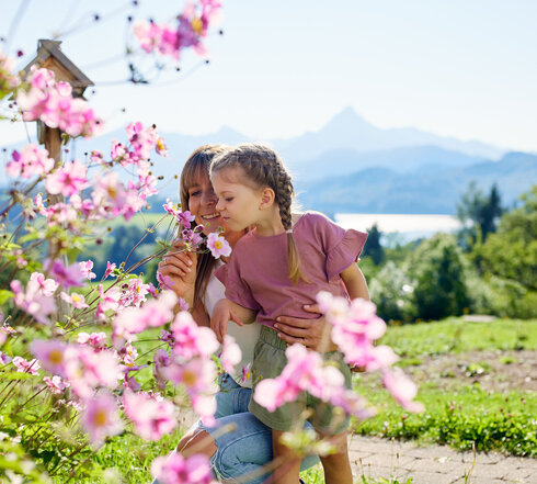 Die Frau und das Kind stehen in einem Garten, der voll von blühenden Pflanzen ist. Die Frau hält einen Strauß Blumen in der Hand und schneidet eine Blume. Das Kind, das auf dem Rücken der Frau sitzt, schaut in die Blumen. Beide sind in einem Moment der Natur zugewandt und genießen die Schönheit der Blumen.  Der Garten liegt in einer hügeligen Landschaft, die von einer Bergkette umgeben ist. Die Blumen sind in verschiedenen Farben, darunter auch in Pink, was die Atmosphäre des Gartens beeinflusst.  Die Frau und das Kind sind in einem Moment der Natur zugewandt und genießen die Schönheit der Blumen. Der Garten liegt in einer hügeligen Landschaft, die von einer Bergkette umgeben ist. Die Blumen sind in verschiedenen Farben, darunter auch in Pink, was die Atmosphäre des Gartens beeinflusst.