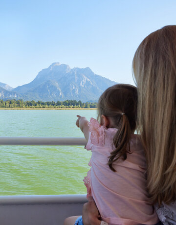 Das Bild fängt eine friedliche Szene von zwei Menschen ein, die mit Kajaks auf einem See unterwegs sind. Die Kajakfahrer tragen Schwimmwesten und paddeln in einem roten Kajak. Der See ist von majestätischen Bergen umgeben, was der Szene ein Abenteuergefühl verleiht. Der Himmel darüber ist ein klarer Blau, was auf einen schönen Tag für Outdoor-Aktivitäten hindeutet. Die Gesamtatmosphäre des Bildes ist eine von Ruhe und Naturgenuss.