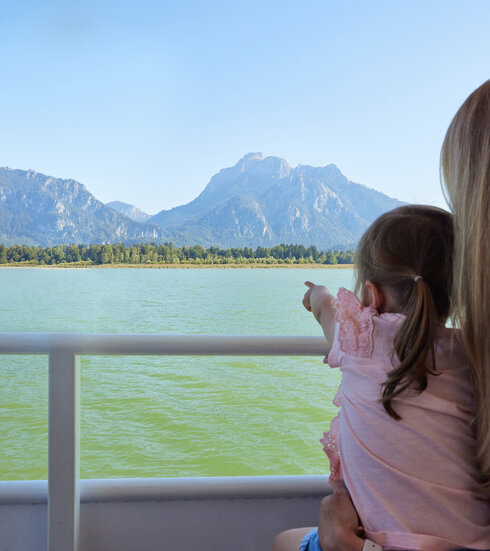 Das Bild fängt eine friedliche Szene von zwei Menschen ein, die mit Kajaks auf einem See unterwegs sind. Die Kajakfahrer tragen Schwimmwesten und paddeln in einem roten Kajak. Der See ist von majestätischen Bergen umgeben, was der Szene ein Abenteuergefühl verleiht. Der Himmel darüber ist ein klarer Blau, was auf einen schönen Tag für Outdoor-Aktivitäten hindeutet. Die Gesamtatmosphäre des Bildes ist eine von Ruhe und Naturgenuss.