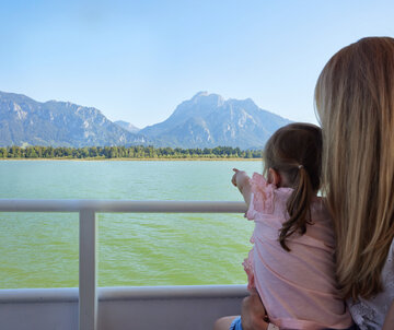 Das Bild fängt eine friedliche Szene von zwei Menschen ein, die mit Kajaks auf einem See unterwegs sind. Die Kajakfahrer tragen Schwimmwesten und paddeln in einem roten Kajak. Der See ist von majestätischen Bergen umgeben, was der Szene ein Abenteuergefühl verleiht. Der Himmel darüber ist ein klarer Blau, was auf einen schönen Tag für Outdoor-Aktivitäten hindeutet. Die Gesamtatmosphäre des Bildes ist eine von Ruhe und Naturgenuss.