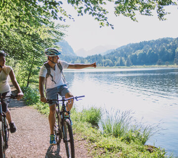Das Bild zeigt einen Mann und eine Frau beim Fahrradfahren. Sie fahren auf E-Bikes, tragen Freizeitkleidung und Helme. Sie fahren auf den Betrachter zu auf einem unbefestigten Uferweg, der auf der rechtenlinken Seite an einen Wald und auf der rechten Seite an einen See grenzt. Die Sonne scheint.