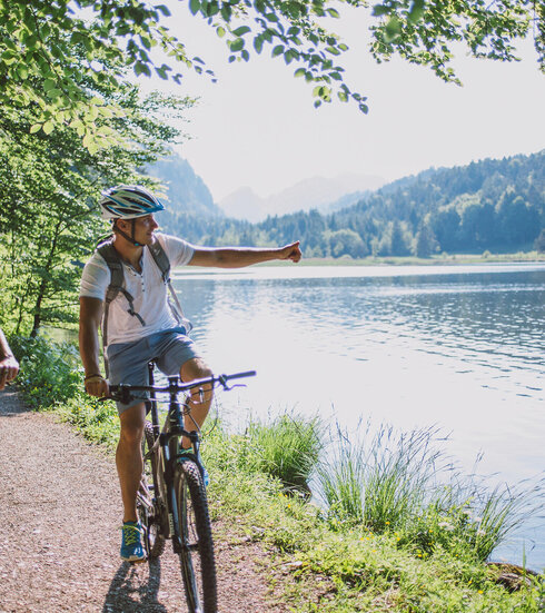 Das Bild zeigt einen Mann und eine Frau beim Fahrradfahren. Sie fahren auf E-Bikes, tragen Freizeitkleidung und Helme. Sie fahren auf den Betrachter zu auf einem unbefestigten Uferweg, der auf der rechtenlinken Seite an einen Wald und auf der rechten Seite an einen See grenzt. Die Sonne scheint.