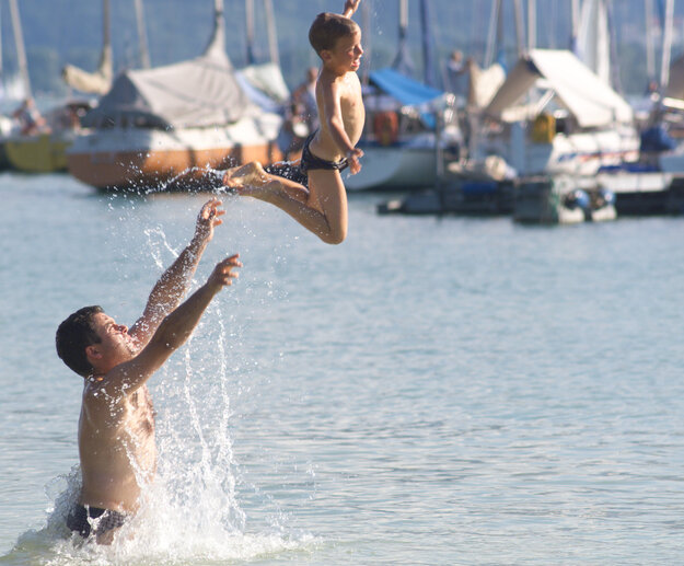 Das Bild zeigt einen Mann und einen Jungen, die im Wasser miteinander toben, spielen und plantschen. Der Mann steht im hüfthohen Wasser. Er wirf den Jungen hoch in die Luft.  Die Sonne scheint. Der Himmel ist blau. Im Hintergrund liegen zahlreiche Segelboote an einem Steg.