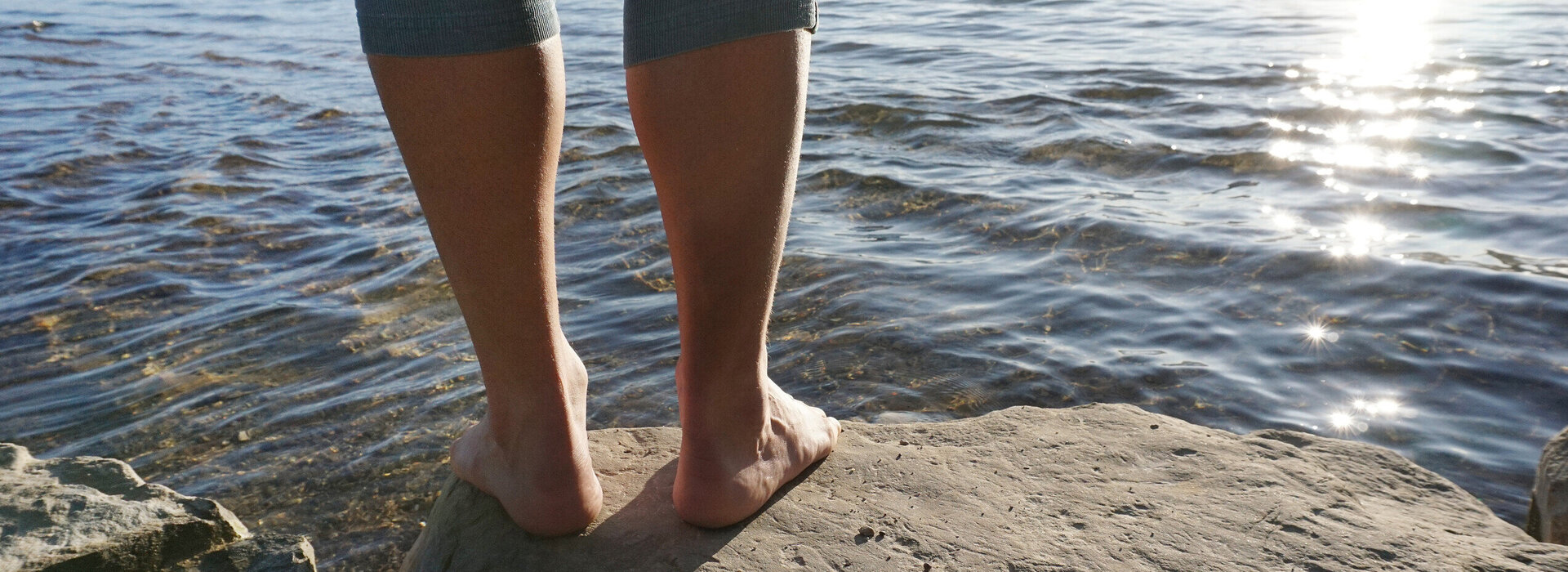 Das Bild zeigt eine friedliche Szene eines Menschen, der auf einem Felsen vor dem Weißensee steht. Es sind lediglich seine Beine zu sehen. Der Mensch trägt eine graublaue Hose, die das Bein hochgeschoben ist, und ist barfuß. Im Wasser kräuseln sich kleine Wellen und die hell aufgehende Sonne glitzert darin. Im Hintergrund sind Berggipfel der Ammergauer Alpen zu sehen.