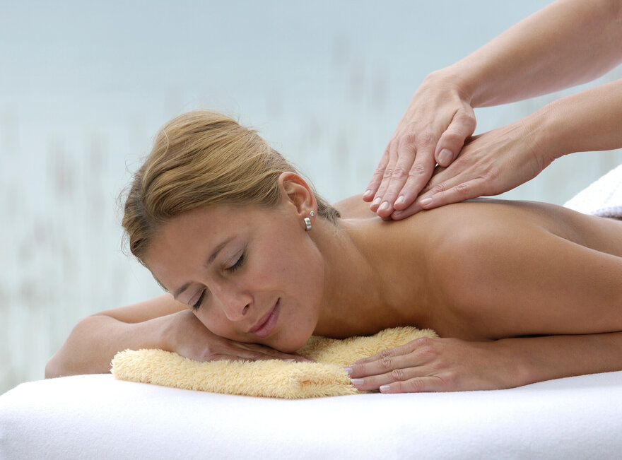 Zu sehen ist eine junge Frau, die oberkörperfrei in Bauchlage auf einer Liege liegt. Ihr Kopf ist auf ein gelbes, zusammengelegtes Handtuch gebettet. Sie lächelt. Vom rechten Bildrand kommend sind zwei weitere Hände zu sehen, die der Frau den Rücken massieren.