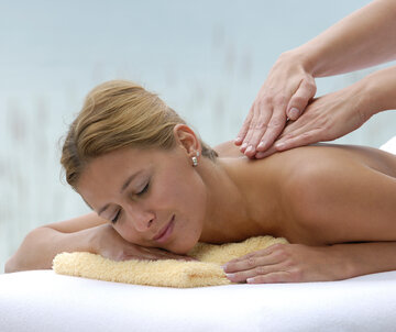 Zu sehen ist eine junge Frau, die oberkörperfrei in Bauchlage auf einer Liege liegt. Ihr Kopf ist auf ein gelbes, zusammengelegtes Handtuch gebettet. Sie lächelt. Vom rechten Bildrand kommend sind zwei weitere Hände zu sehen, die der Frau den Rücken massieren.