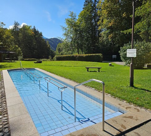 Das Bild zeigt eine grüne Wiese, auf der vereinzelt Liegen und Bänke stehen. Eine grüne Hecke rahmt einen Kiesplatz ein. Im Hintergrund beginnt der Laubwald. Vor dem Laubwald steht ein hölzerner Pavillon. Im Vordergrund ist ein blau, gefliestes Tretbecken zu sehen. In der Mitte des Tretbeckens ist ein silbernes Metallgeländer angebracht. Die Sonne scheint. Der Himmel ist blau.
