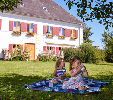 Das Bild zeigt eine ruhige Szene von zwei jungen Mädchen, die einen sonnigen Tag in einem üppigen grünen Garten genießen. Sie sitzen auf einem lebhaft blauen Tuch, das auf dem Gras ausgebreitet ist. Das Mädchen auf der linken Seite ist vertieft in ein Buch, ihre Aufmerksamkeit ganz gefangen von den Seiten. Das Mädchen auf der rechten Seite hingegen trinkt aus einer Tasse einen erfrischenden Getränk.  Der Garten ist von einem weißen Haus im Hintergrund geschmückt, dessen rote Fensterläden eine Farbtupfer zur Szene hinzufügen. Das Haus ist eingebettet in einer Anordnung von Bäumen und Sträuchern, die eine malerische Kulisse für das Outdoor-Abenteuer der Mädchen schaffen. Die Gesamtatmosphäre des Bildes ist eine von Ruhe und Genuss, während die Mädchen die Schönheit der Natur in vollen Zügen genießen und ihre Lieblingsbeschäftigungen nachgehen.