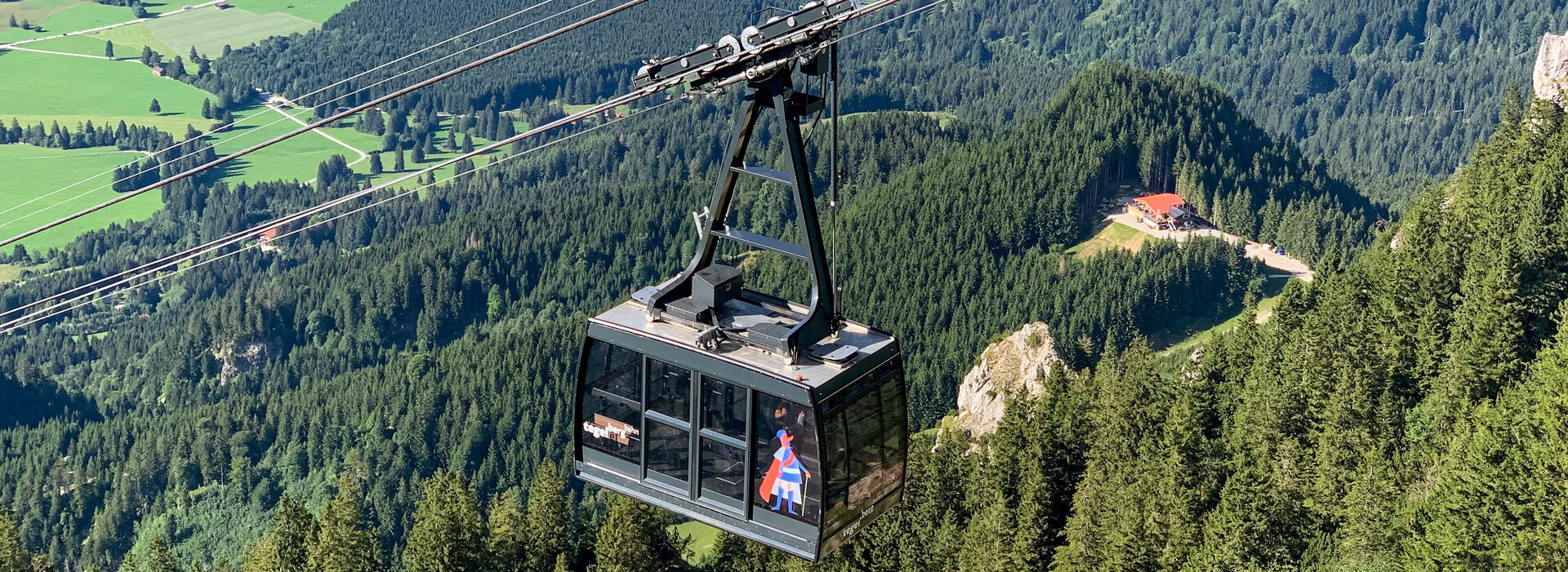 Das Bild zeigt die an einem Seil hängende Kabine einer Bergbahn. Im Tal erblickt man zwei blaue Seen, Wiesen und Wälder. Die Sonne scheint. 