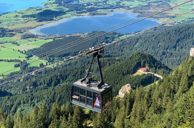 Das Bild zeigt die an einem Seil hängende Kabine einer Bergbahn. Im Tal erblickt man zwei blaue Seen, Wiesen und Wälder. Die Sonne scheint. 