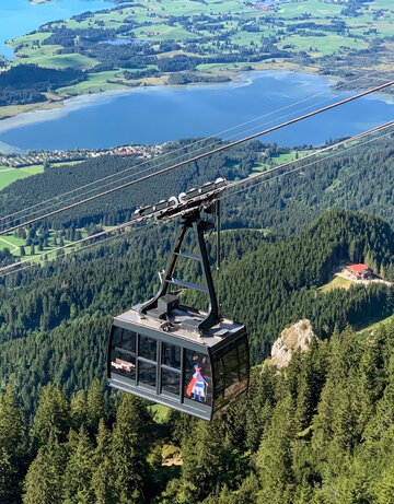 Das Bild zeigt die an einem Seil hängende Kabine einer Bergbahn. Im Tal erblickt man zwei blaue Seen, Wiesen und Wälder. Die Sonne scheint. 