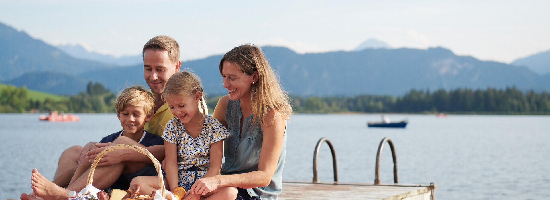 Das Bild fängt einen friedlichen Familienmoment am Ufer eines Sees ein. Eine Familie bestehend aus einem Mann, einer Frau und zwei Kindern sitzt auf einer Holzdiele. Der Mann sitzt auf der linken Seite, die Frau sitzt in der Mitte und die zwei Kinder sitzen auf der rechten Seite. Sie alle lächeln und schauen zur Kamera, wodurch eine warme und glückliche Atmosphäre geschaffen wird.  Die Familie wird von der Natur umgeben, mit dem See und den Bergen im Hintergrund. Die Holzdiele, auf welcher die Familie sitzt, ist rot und weiß, was eine Farbtupfer im Bild schafft.  Im Vordergrund befindet sich ein Korb voller Essen, was darauf hindeutet, dass die Familie vielleicht ein Picknick hat. Die Gesamtszene ist eine der Entspannung und Freude an der Natur.