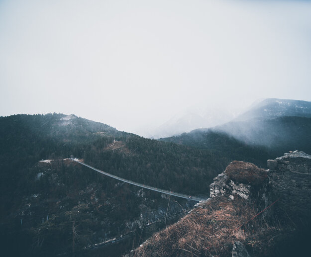 Das Bild zeigt eine atemberaubende Landschaft mit Gebirgen. Die Szene wird von einer windenden Straße dominiert, die sich wie eine Schlange durch das Gelände schlängelt und in der Ferne verschwindet. Die Straße, ein Faden aus grauem Asphalt, ist von saftig grünen Bäumen und Sträuchern flankiert, deren leuchtende Farben schön im Kontrast zum Grau der Straße stehen. Der Himmel darüber ist ein helles Blau, durchzogen von zarten Wolken, die der Szene Tiefe und Weite verleihen. Die Perspektive des Bildes ist eine hohe Aufsicht, die auf die Straße und die umgebende Landschaft hinabschaut. Diese Perspektive erlaubt eine umfassende Sicht auf das Gebiet und zeigt den windenden Pfad der Straße und die dichte Vegetation, die sie säumt. Die Gesamtkomposition des Bildes erzeugt ein Gefühl der Ruhe und Ehrfurcht und fängt die natürliche Schönheit der Gebirgslage ein.