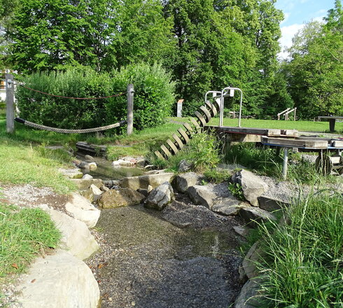 Das Bild zeigt einen kleinen Bach, an dem unterschiedliche Spiegelgeräte angebracht sind. Über den Bach führt ein Balanciertau. Eine übergroße Schraubvorrichtung liegt mit der Spitze im Wasser. Ein Wassertisch ist neben dem Bach aufgestellt. Der Platz ist von hohen Laubbäumen eingerahmt. Der Himmel ist blau. Die Sonne scheint. 