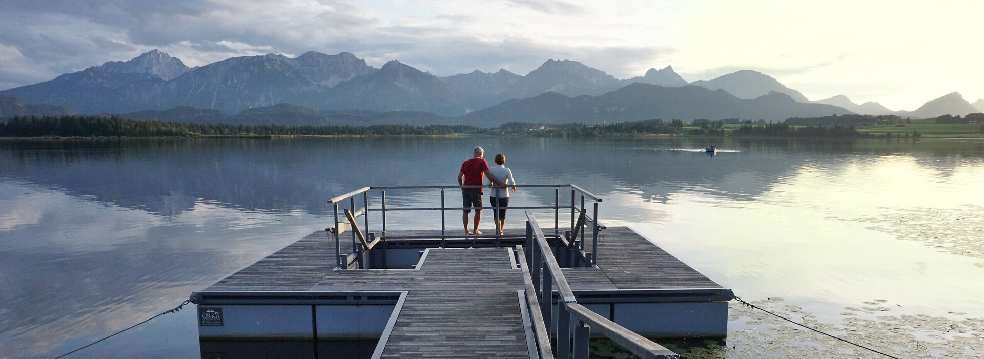 Das Bild zeigt einen großen Steg, der in den Hopfensee ragt. Auf dem See schwimmen Seerosenblätter. Am Ende des Stegs wird öffnet sich eine Plattform, in deren Mitte ein Kneipptretbecken eingelassen ist. Auf der Kneippinsel stehen zwei Menschen mit dem Rücken an ein Geländer gelehnt und blicken in die Ferne. Der Himmel ist in ein ruhiges Abendlicht getaucht und im Hintergrund erhebt sich die Bergkette der Allgäuer Alpen.