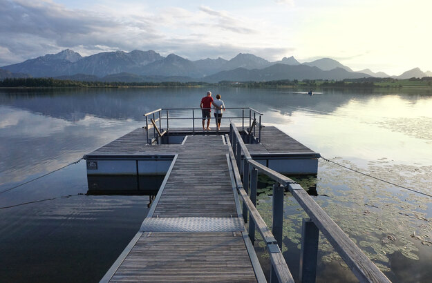 Das Bild zeigt einen großen Steg, der in den Hopfensee ragt. Auf dem See schwimmen Seerosenblätter. Am Ende des Stegs wird öffnet sich eine Plattform, in deren Mitte ein Kneipptretbecken eingelassen ist. Auf der Kneippinsel stehen zwei Menschen mit dem Rücken an ein Geländer gelehnt und blicken in die Ferne. Der Himmel ist in ein ruhiges Abendlicht getaucht und im Hintergrund erhebt sich die Bergkette der Allgäuer Alpen.