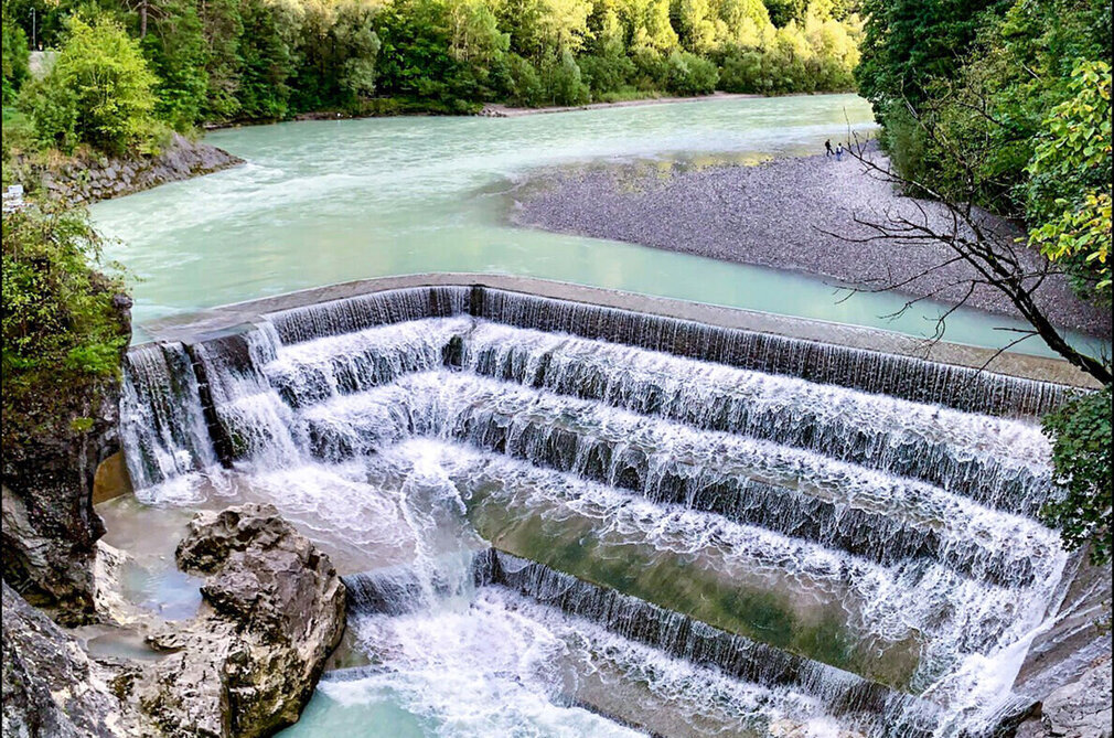 Das Bild zeigt einen atemberaubenden Blick auf einen Wasserfall – den Lechfall bei Füssen, der sich über eine felsige Klippe hinunterstürzt. Der Wasserfall, mit seinem weißen Schaum, ist der Hauptakzent im Bild. Er ist von üppig grünen Bäumen und Büschen umgeben, die der Szene eine Prise der Naturpracht verleihen. Der Wasserfall liegt in einem Tal, durch das der Fluss Lech fließt. Der klare blaue Himmel über ihm ist von fluffigen, weißen Wolken durchzogen. Die Perspektive des Bildes ist aus einer hohen Position, was eine Panoramaansicht des Wasserfalls und seiner Umgebung bietet.