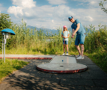 Das Bild zeigt eine friedliche Szene eines Mannes und eines jungen Mädchens, die Golf spielen auf einer grünen Wiese. Der Mann, in einem blauen Hemd und blauen Shorts gekleidet, ist dabei, seinen Golfschläger zu schwingen und auf den Golfball zu zielen, der auf dem Boden liegt. Das Mädchen, das einen pinken Hut trägt, steht in der Nähe und beobachtet aufmerksam den Schwung des Mannes. Die Wiese, auf der sie sich befinden, ist üppig und grün, mit hohen Gräsern, die sanft im Wind wehen. In der Ferne erstreckt sich ein majestätisches Gebirge über den Horizont und verleiht der Szene eine beruhigende Ruhe. Der Himmel ist ein klarer blauer Farbton, mit flauschigen weißen Wolken verstreut. Das Bild vermittelt ein Gefühl von Leichtigkeit und Freude an der Natur.