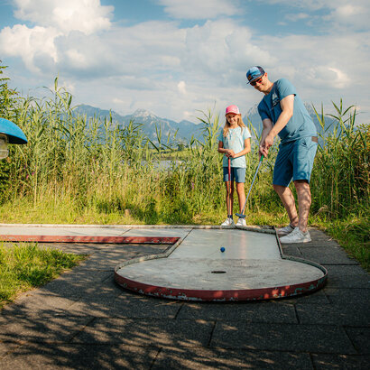 Das Bild zeigt eine friedliche Szene eines Mannes und eines jungen Mädchens, die Golf spielen auf einer grünen Wiese. Der Mann, in einem blauen Hemd und blauen Shorts gekleidet, ist dabei, seinen Golfschläger zu schwingen und auf den Golfball zu zielen, der auf dem Boden liegt. Das Mädchen, das einen pinken Hut trägt, steht in der Nähe und beobachtet aufmerksam den Schwung des Mannes. Die Wiese, auf der sie sich befinden, ist üppig und grün, mit hohen Gräsern, die sanft im Wind wehen. In der Ferne erstreckt sich ein majestätisches Gebirge über den Horizont und verleiht der Szene eine beruhigende Ruhe. Der Himmel ist ein klarer blauer Farbton, mit flauschigen weißen Wolken verstreut. Das Bild vermittelt ein Gefühl von Leichtigkeit und Freude an der Natur.