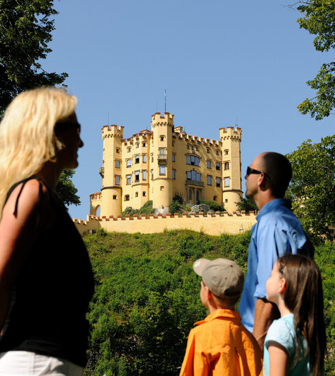 Das Bild zeigt eine friedliche Szene einer Familie bestehend aus einer Frau, einem Mann und zwei Kindern, die vor einem Schloss stehen. Die Frau, in einem schwarzen Oberteil gekleidet, steht links im Bild. Sie schaut in Richtung rechts, ihr Blick folgt dem Mann und den zwei Kindern. Der Mann trägt ein blaues Hemd und steht in der Mitte des Bildes, flankiert von den beiden Kindern, das eine in einem orangefarbenen Hemd und das andere in einem blauen Hemd. Das Schloss, das farblich in einem leuchtenden Gelb gestaltet ist, steht majestätisch im Hintergrund. Die Familie scheint einen Ausflug zu genießen und die Ansichten des Schlosses zu bewundern.