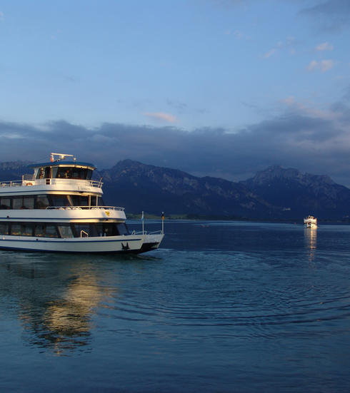 Das Foto zeigt einen See. Auf dem zwei Schiffe schwimmen. Das linke Schiff steht im Vordergrund. Das rechte Schiff kommt auf den Betrachter zu und ist eher klein im Hintergrund. Im Bildhintergrund erheben sich die Berge. Man kann die Schlösser Neuschwanstein und Hohenschwangau davor erahnen. Der Himmel ist blau mit vereinzelten Wolken. Die Sonne geht unter. Es ist Sommer. 