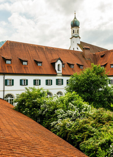 Das Foto zeigt einen Teil es Gebäudekomplexes des Franziskanerklosters. Mit seinen roten Dächern und vielen Fenstern, die von grünen Fensterläden umrahmt sind. Im Hintergrund erhebt sich der Kirchturm der Klosterkirche St. Stephan. Im Vordergrund ist eine Teil des Klostergartens zu sehen, in dem Büsche und Bäume wachsen. Der Himmel ist wolkenverhangen. Es ist Frühling.