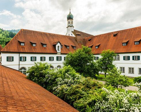 Das Foto zeigt einen Teil es Gebäudekomplexes des Franziskanerklosters. Mit seinen roten Dächern und vielen Fenstern, die von grünen Fensterläden umrahmt sind. Im Hintergrund erhebt sich der Kirchturm der Klosterkirche St. Stephan. Im Vordergrund ist eine Teil des Klostergartens zu sehen, in dem Büsche und Bäume wachsen. Der Himmel ist wolkenverhangen. Es ist Frühling.  