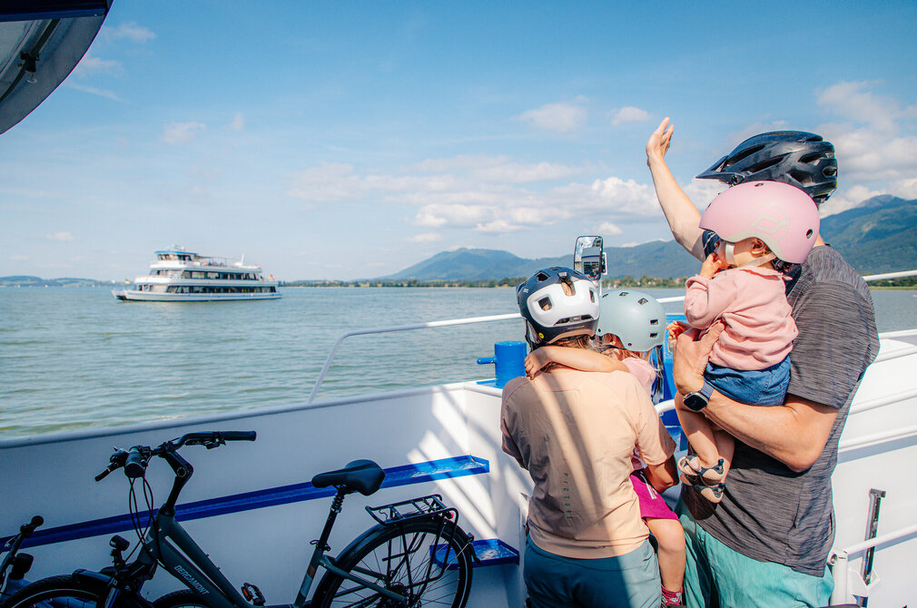 Das Bild zeigt eine idyllische Szene auf einem Boot. Ein Mann, der mit einer rosa Badehaube ausgestattet ist, hält ein Baby in den Armen. Das Baby trägt ebenfalls eine rosa Badehaube, was auf das gemeinsame Interesse an der Sicherheit hinweist. Der Mann hält auch eine rosa Badehaube, was darauf hindeuten könnte, dass er der Vater des Babys ist.  Das Boot, auf dem sie sich befinden, ist weiß und blau und liegt in einem Hafen. Der Hafen wird von Bergen umgeben, was der Landschaftszene etwas Natur verleiht. In der Ferne ist ein großes Boot zu sehen, was darauf hindeuten könnte, dass sie sich in einem beliebten Bereich für das Wassersport befinden.  Der Mann und das Baby sind nicht allein auf dem Boot. Auch ein Fahrrad ist anwesend, was darauf hinweisen könnte, dass sie eine gemütliche Fahrt oder eine Exkursion genießen. Das Fahrrad steht fertig auf dem Boot bereit, sobald sie ihr Ziel erreicht haben.  Insgesamt erzeugt das Bild den Eindruck einer Familie, die einen Tag auf dem Wasser genießt, mit Sicherheit an erster Stelle.